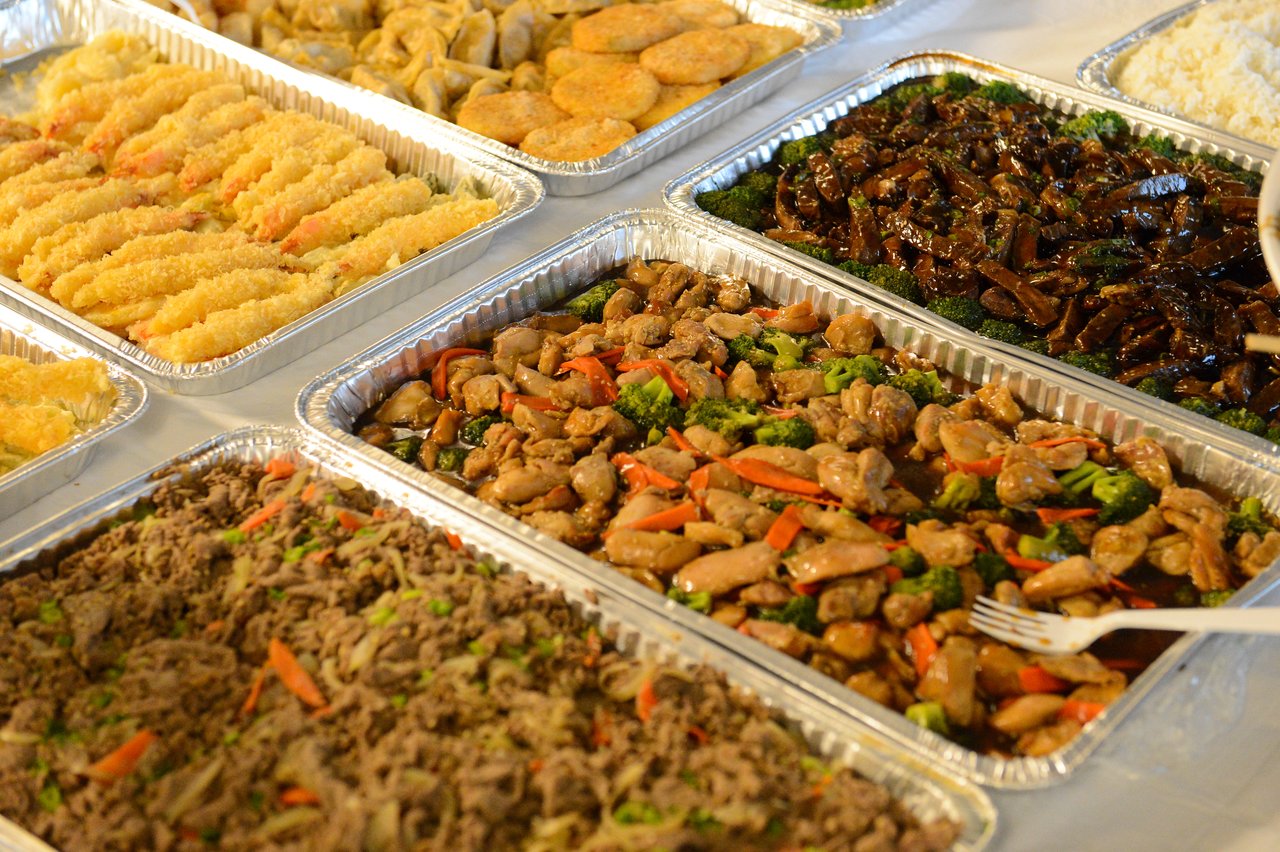 Trays of assorted food, including stir-fried chicken with vegetables, fried shrimp, and other dishes, set up for a buffet.