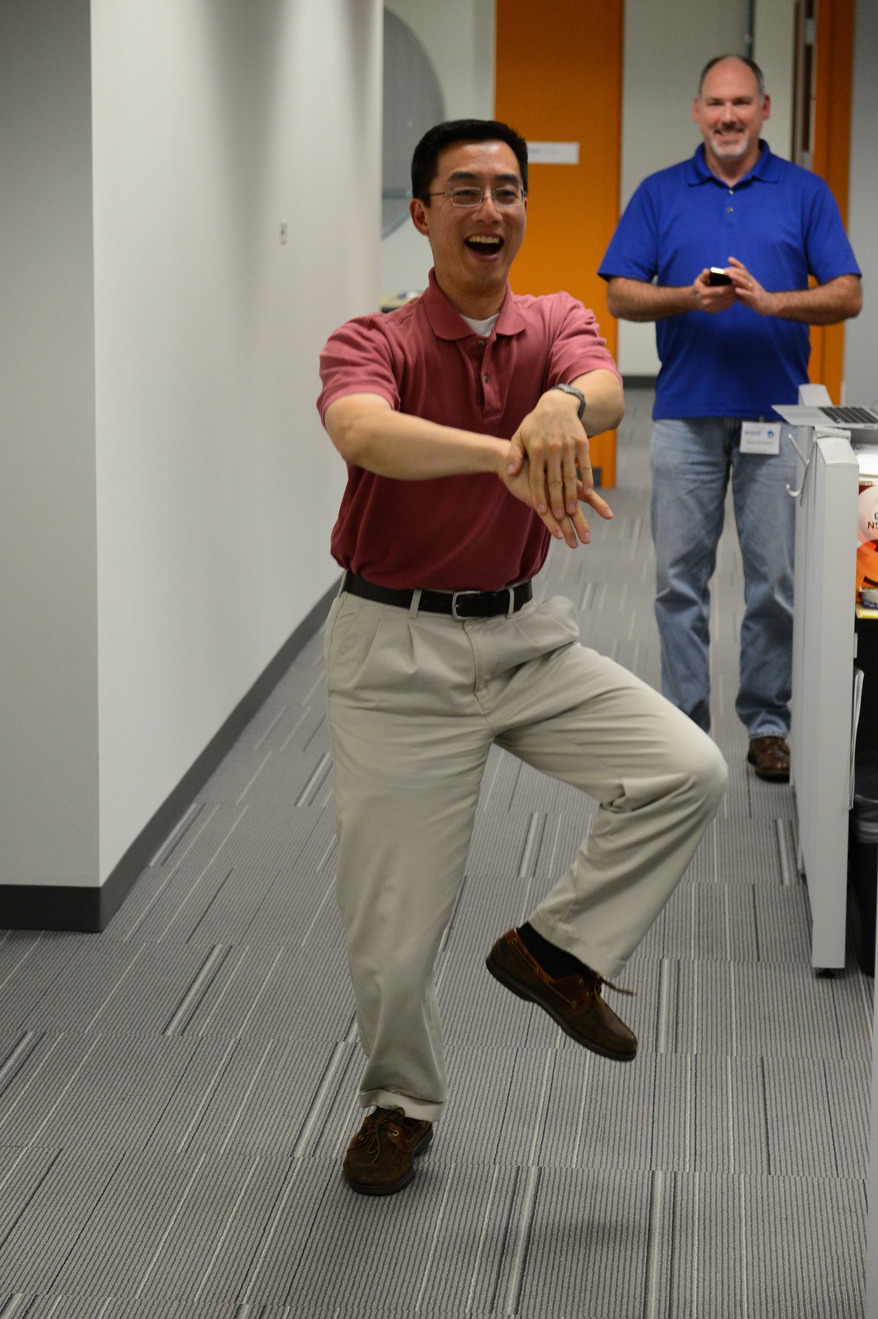 A man in a red shirt playfully dances in an office while another man in blue watches and smiles.