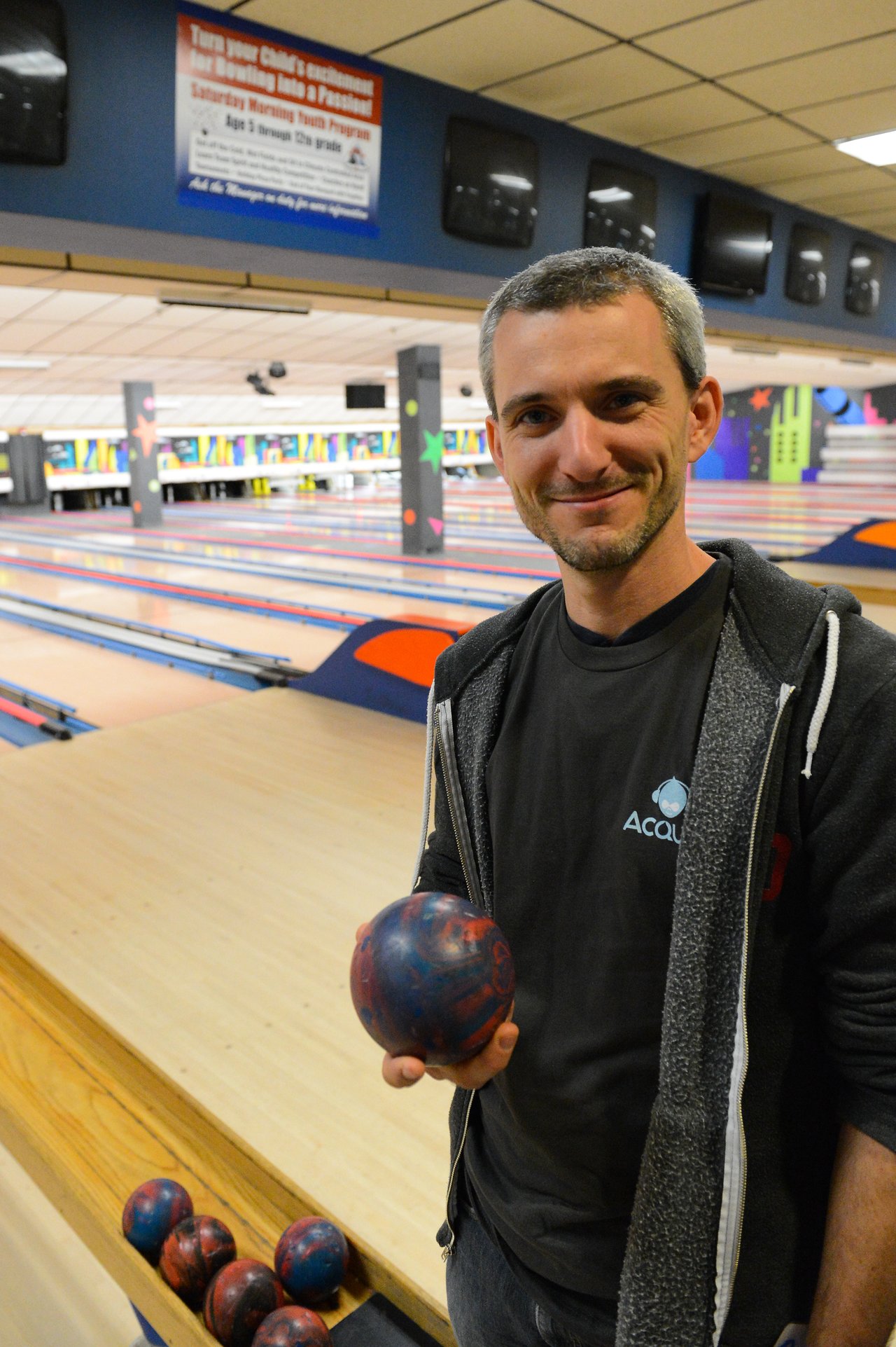 A person wearing an Acquia shirt smiles while holding a bowling ball at a bowling alley.