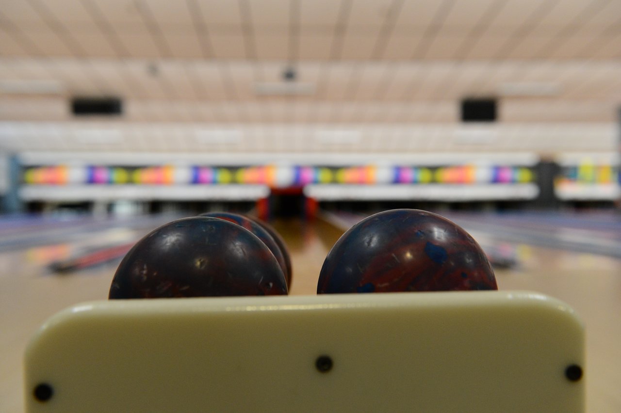 Two bowling balls rest on a rack at a bowling alley, with lanes and pins in the background.