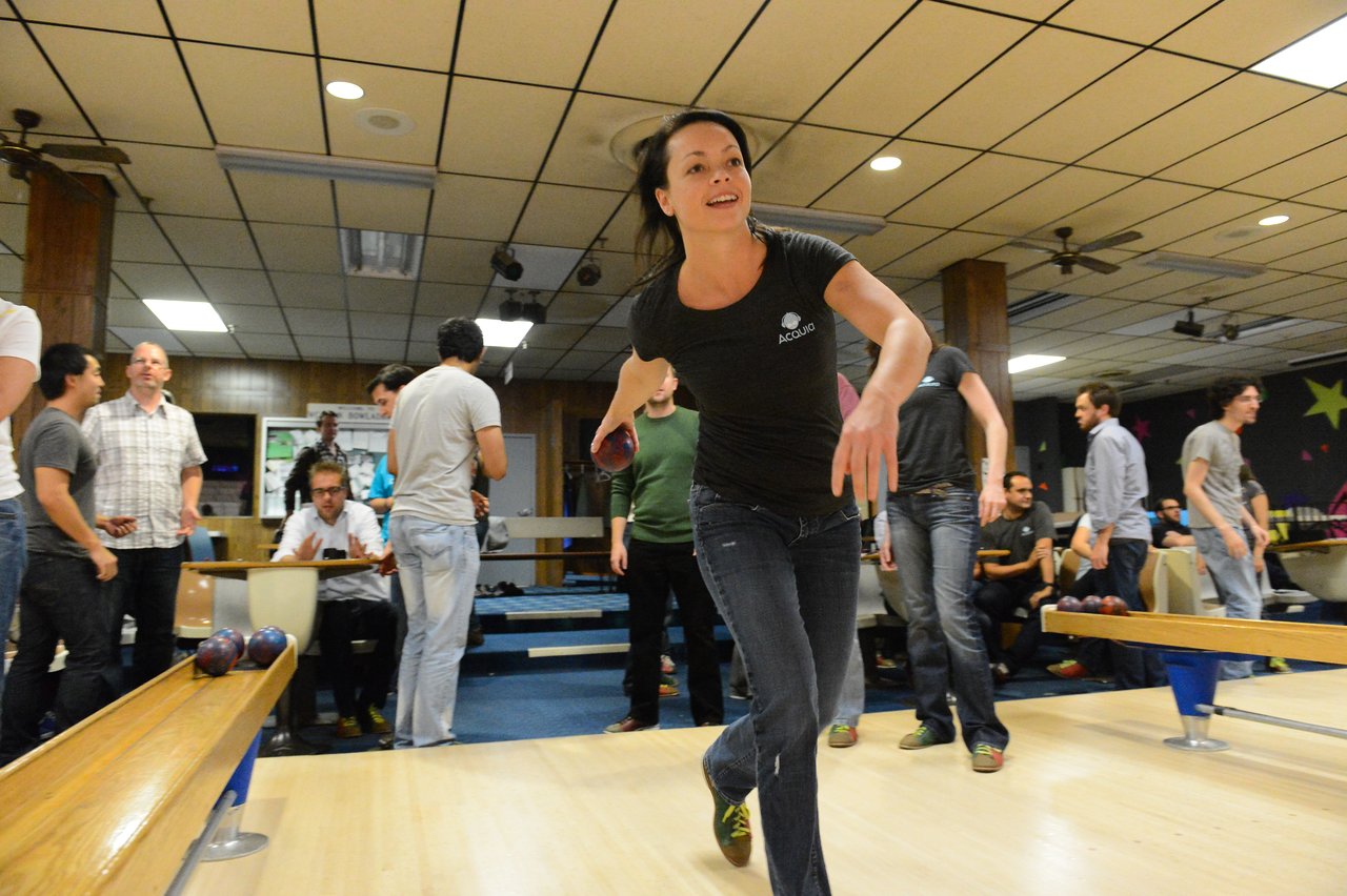 A woman in a black Acquia shirt releases a bowling ball, surrounded by colleagues at a bowling alley.