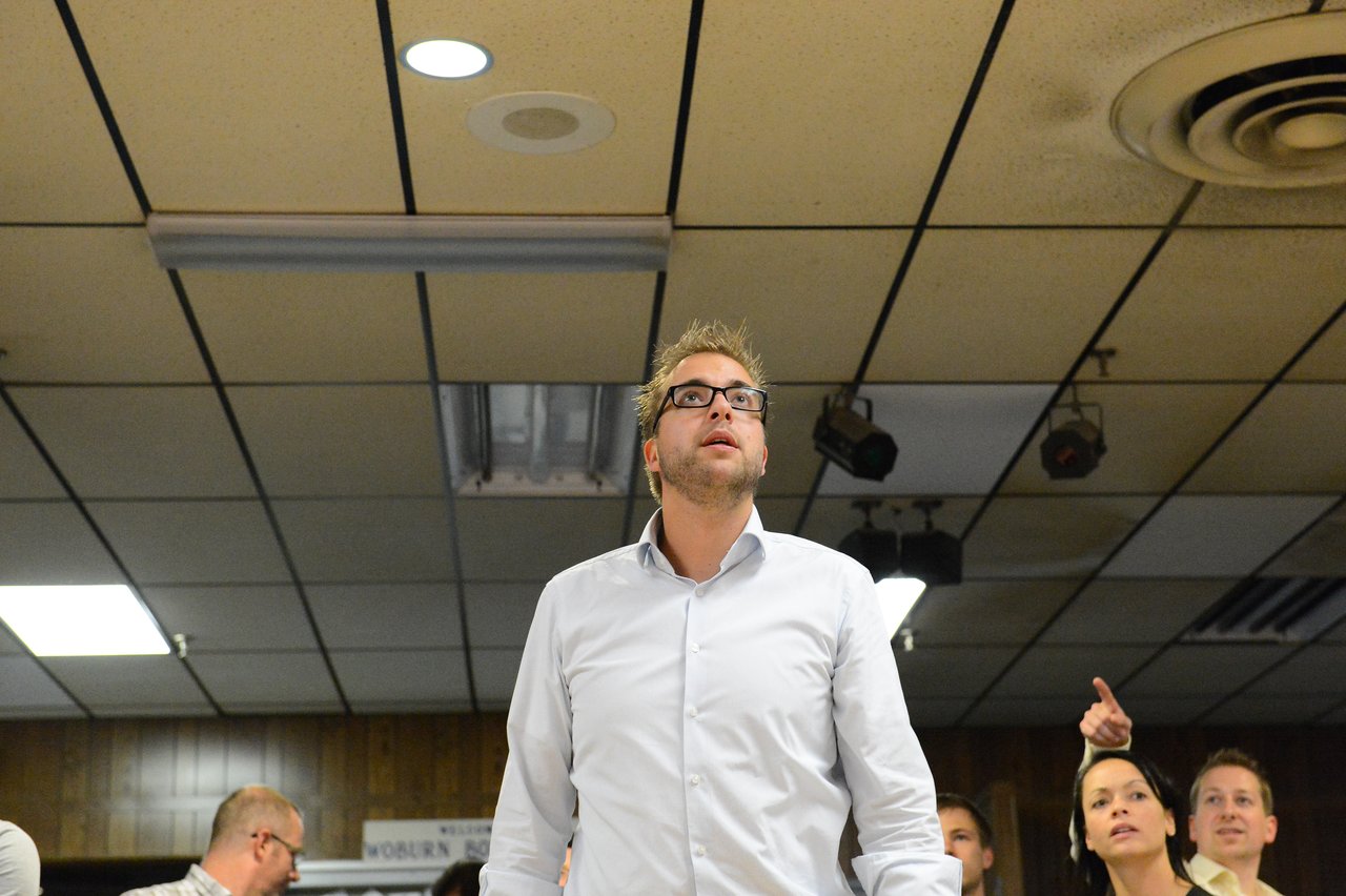 A man in a white shirt and glasses looks up, while others in the background react and point.