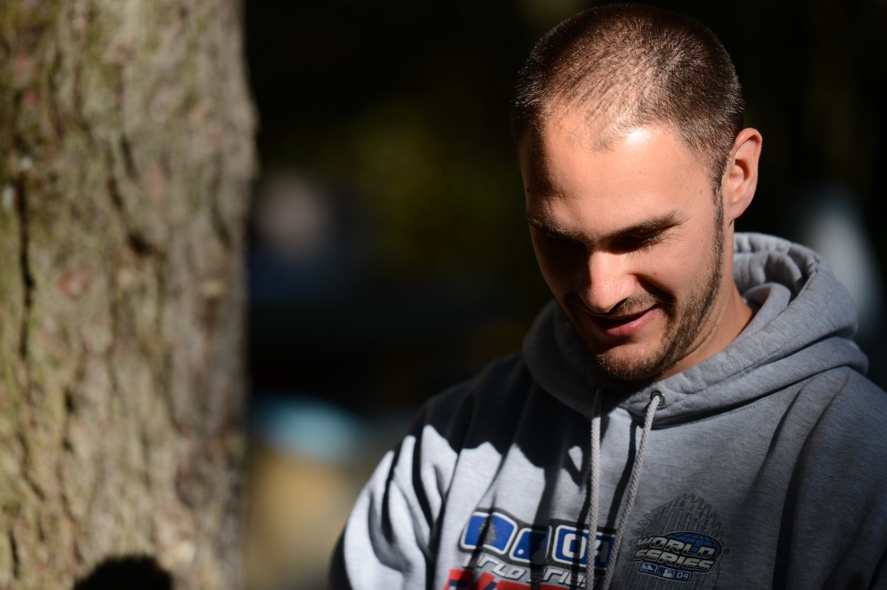 A man in a gray hoodie looks down and smiles while standing outdoors in natural light.