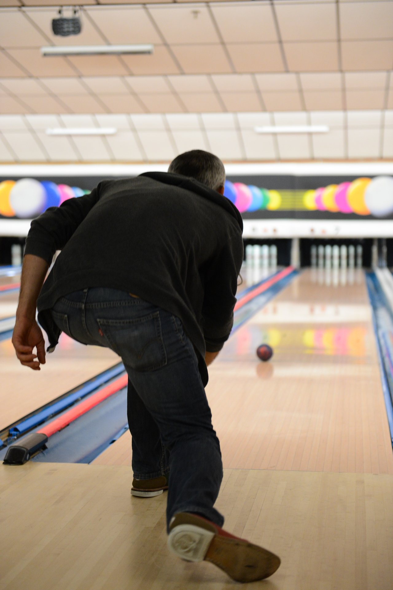 A person in a dark jacket and jeans bowls a ball down a lane at a bowling alley.
