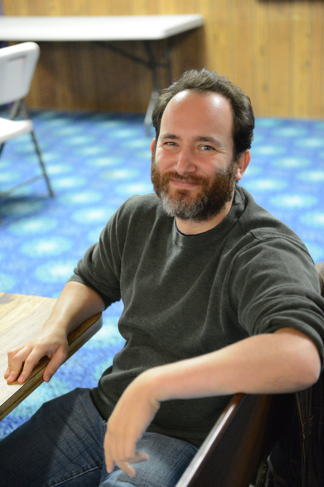 A man with a beard and dark sweater sits at a table, smiling at the camera.