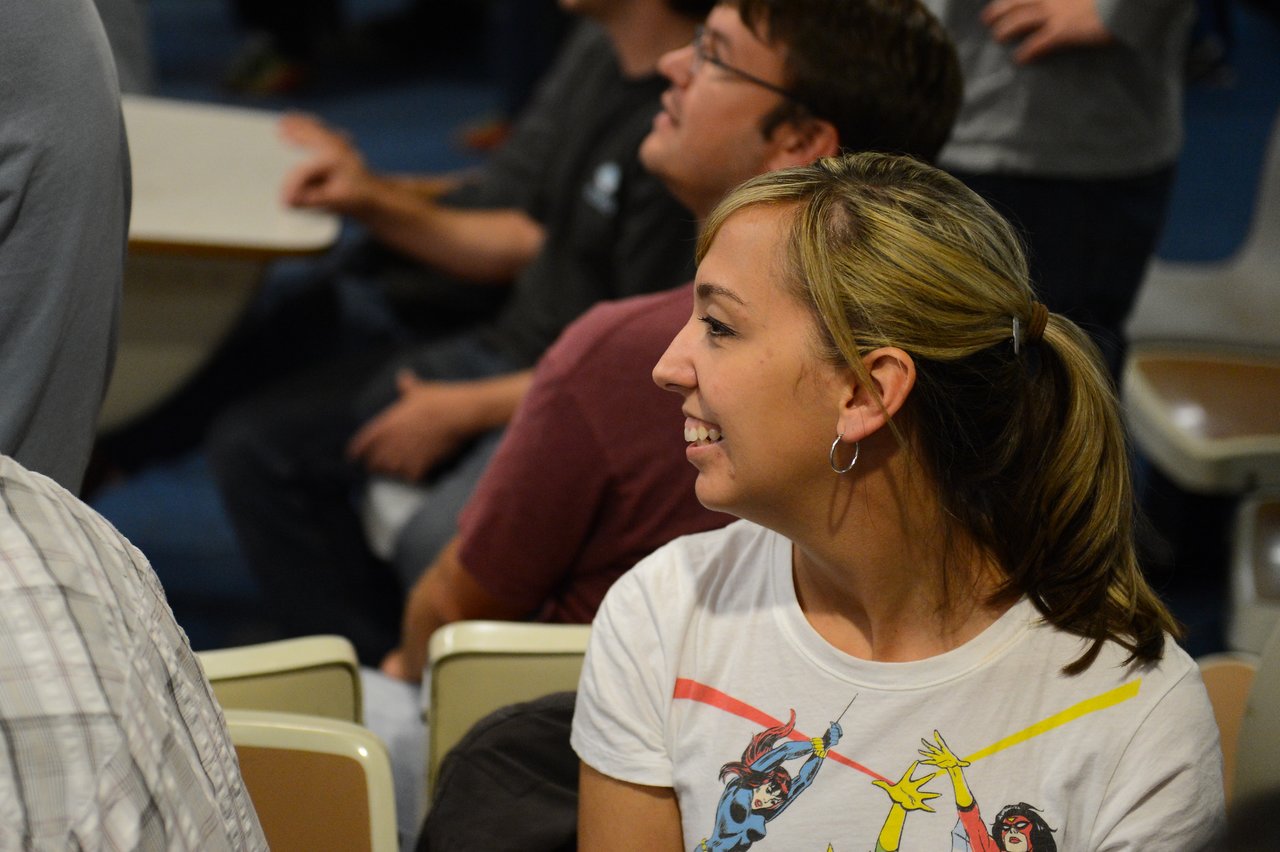 A woman with a ponytail and a graphic t-shirt smiles while looking to the side in a crowded room.