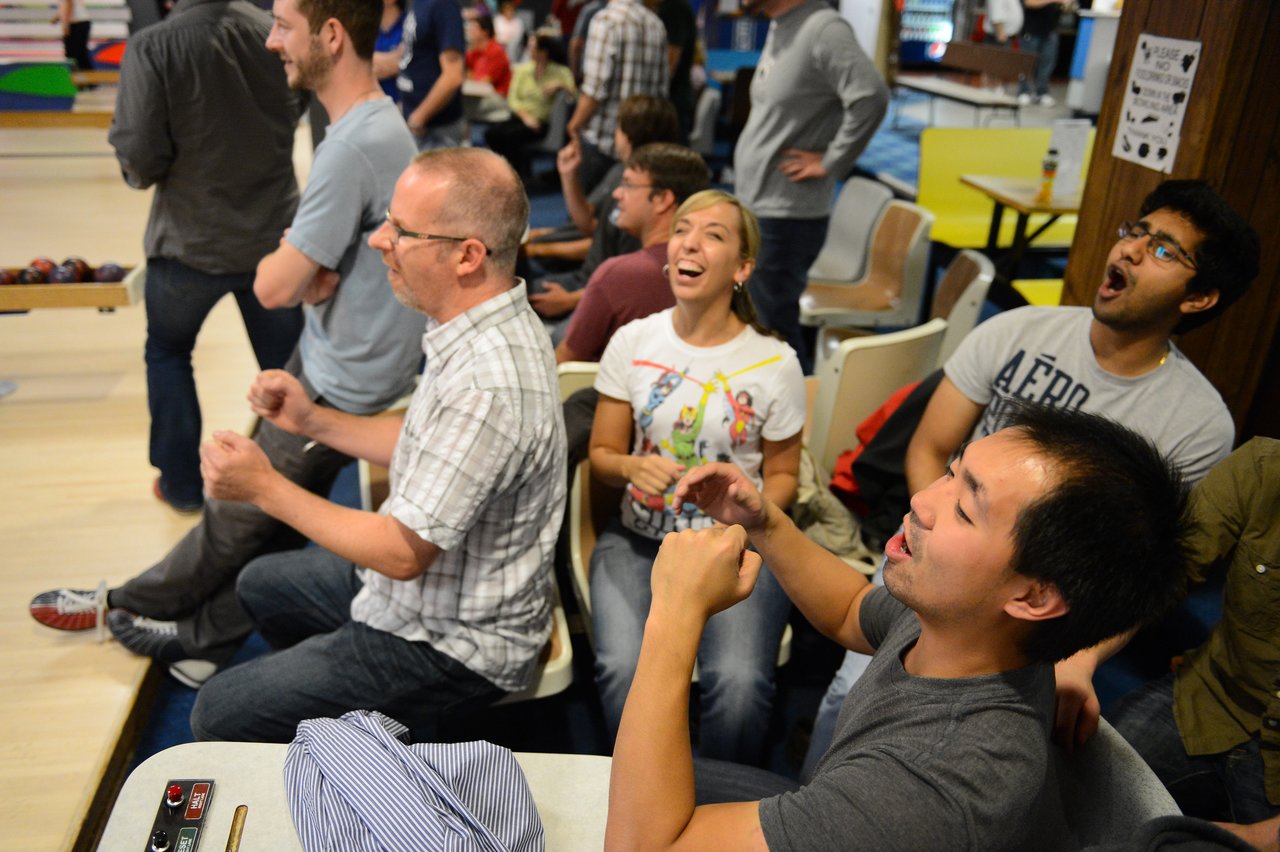 A group of people at a bowling alley react excitedly while watching a game, cheering and gesturing.