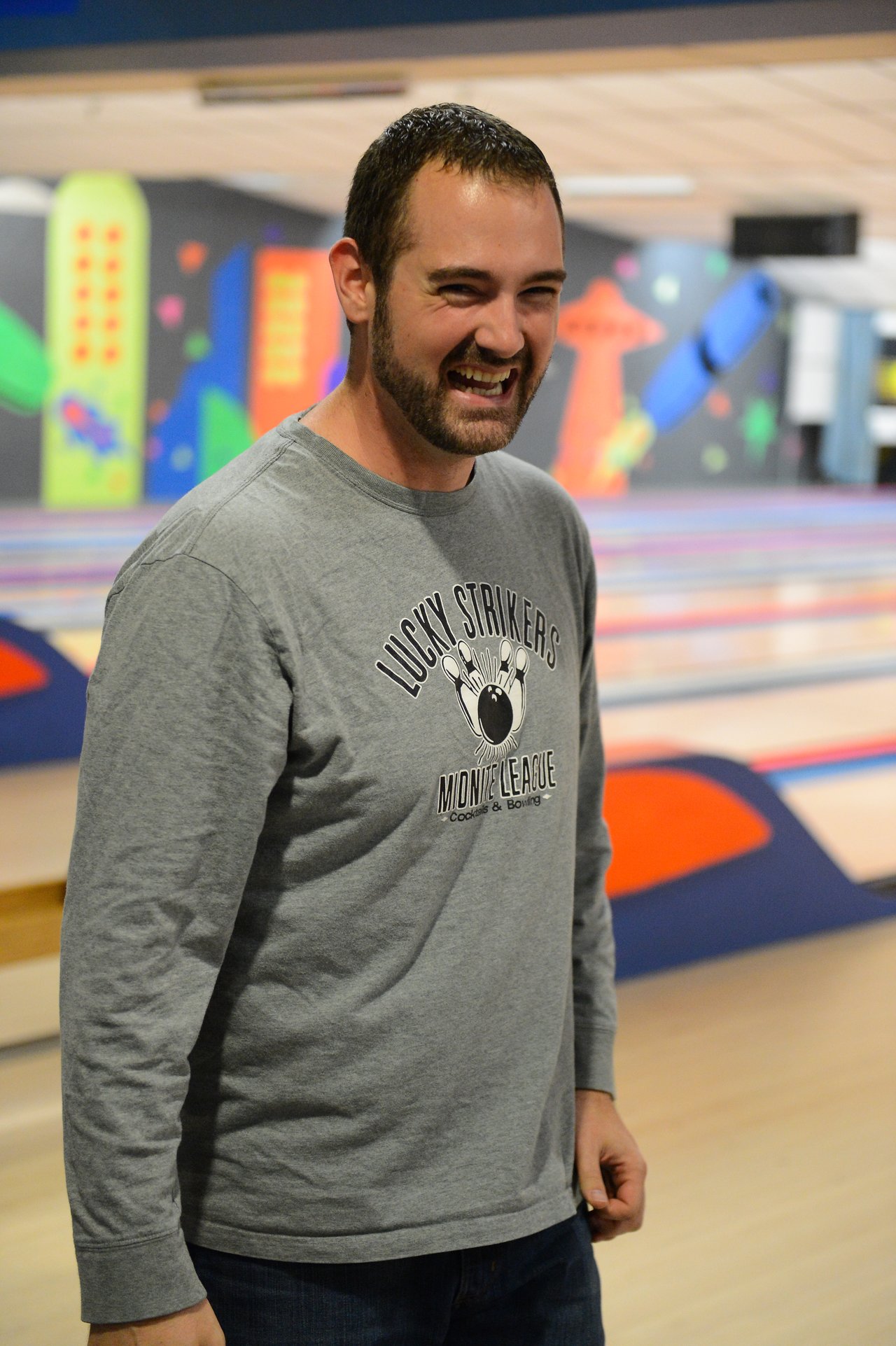 A man in a gray bowling league shirt smiles while standing in a bowling alley.
