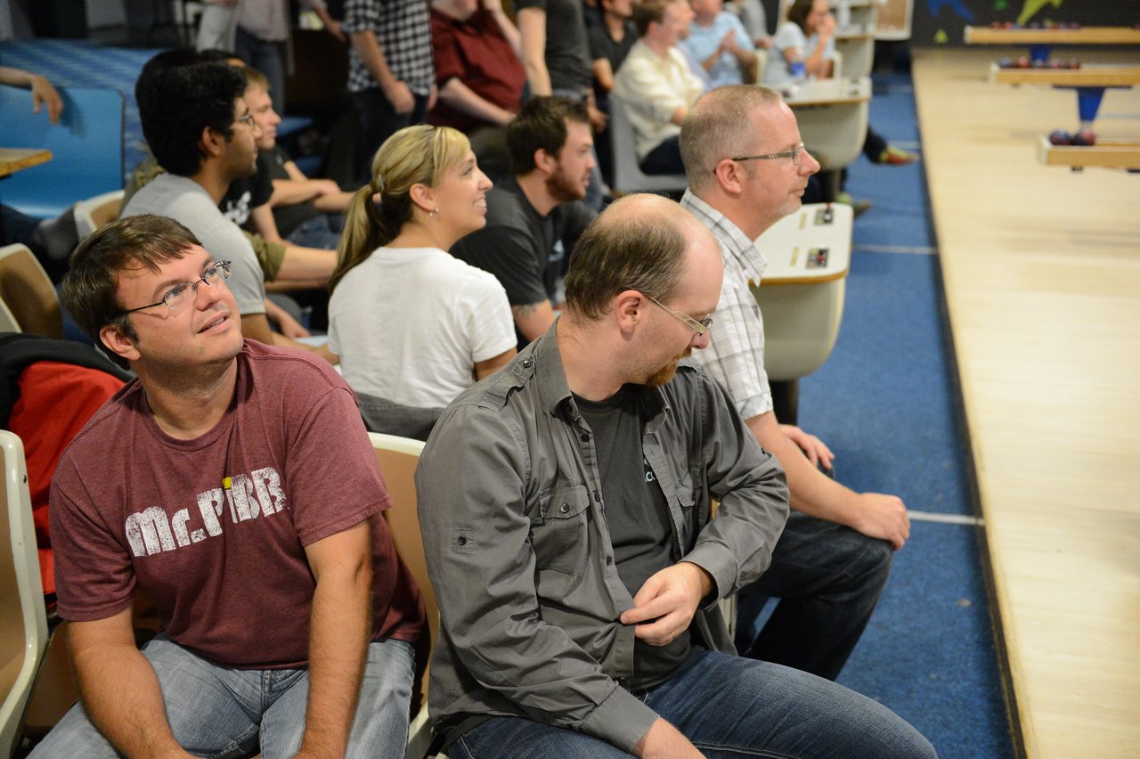 A group of people sits near a bowling lane, watching the game and reacting to the action.