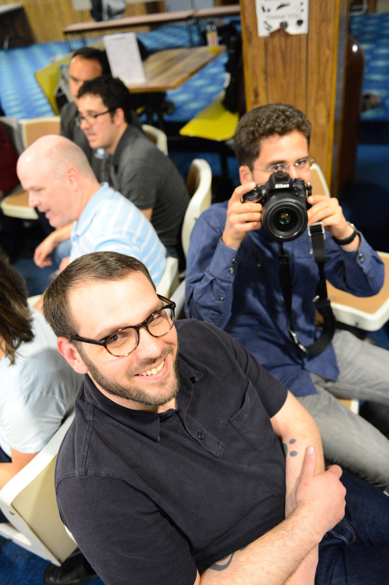 A smiling man in glasses sits in the foreground, while another person behind him holds a camera and takes a photo.