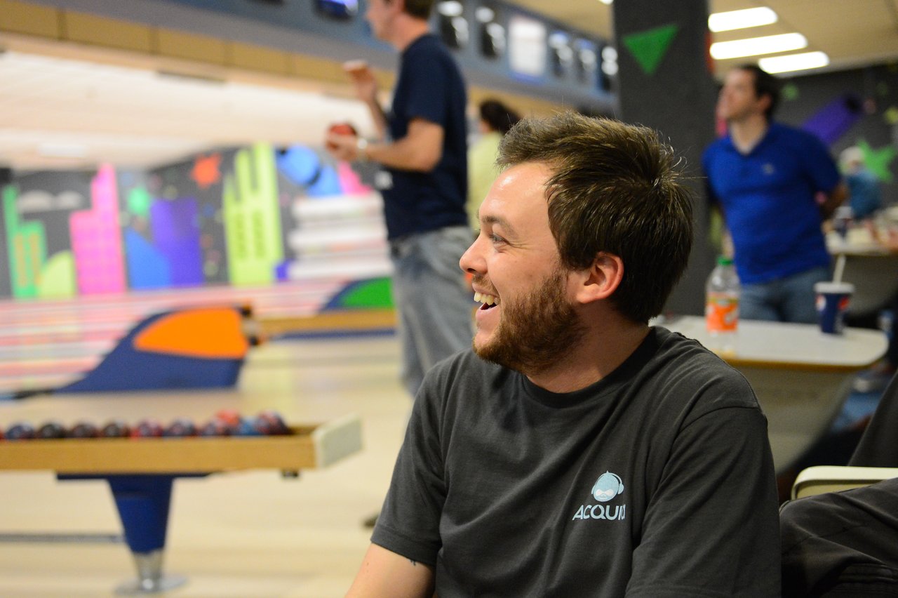 A man wearing an Acquia shirt laughs while sitting at a bowling alley with others in the background.