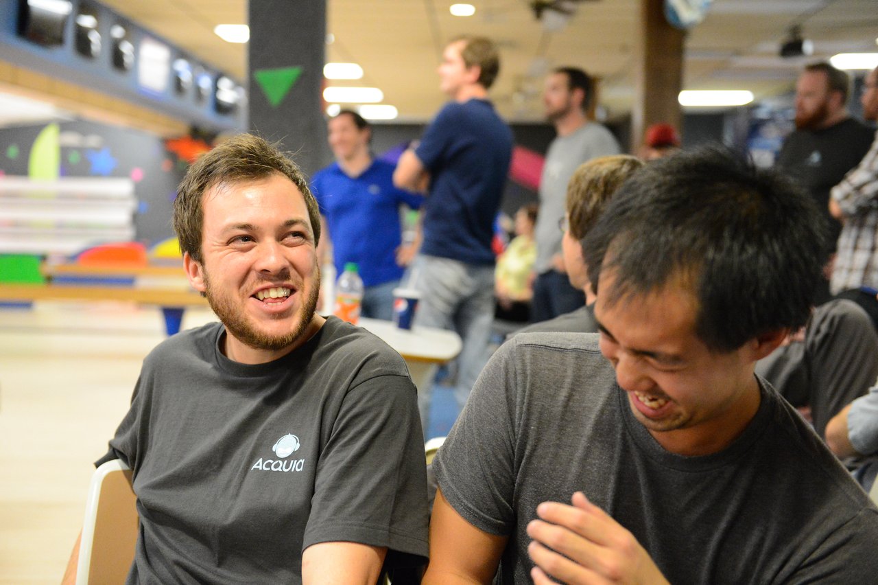 Two people wearing Acquia shirts are sitting and laughing together at a bowling alley during a group event.