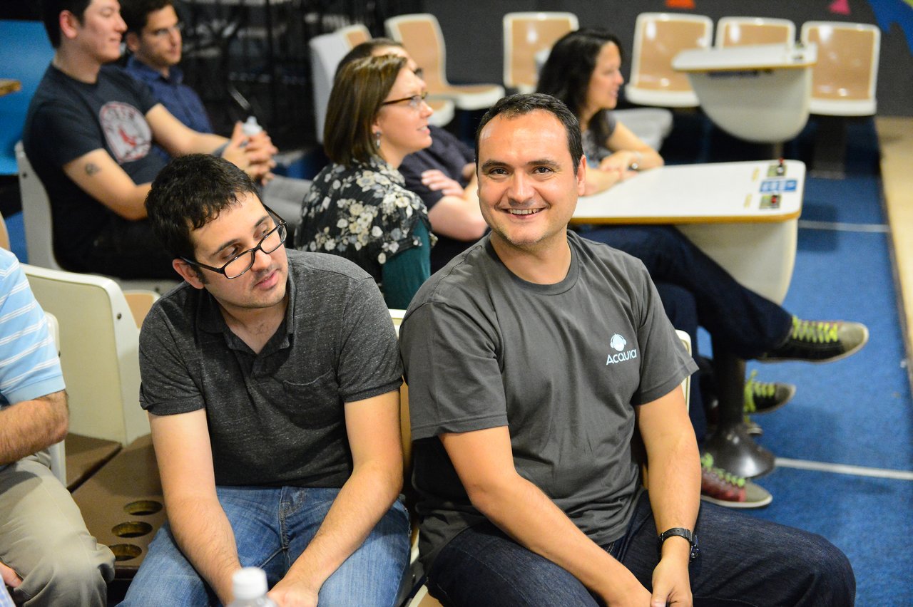 Two men sit close together, one smiling at the camera while the other looks to the side.
