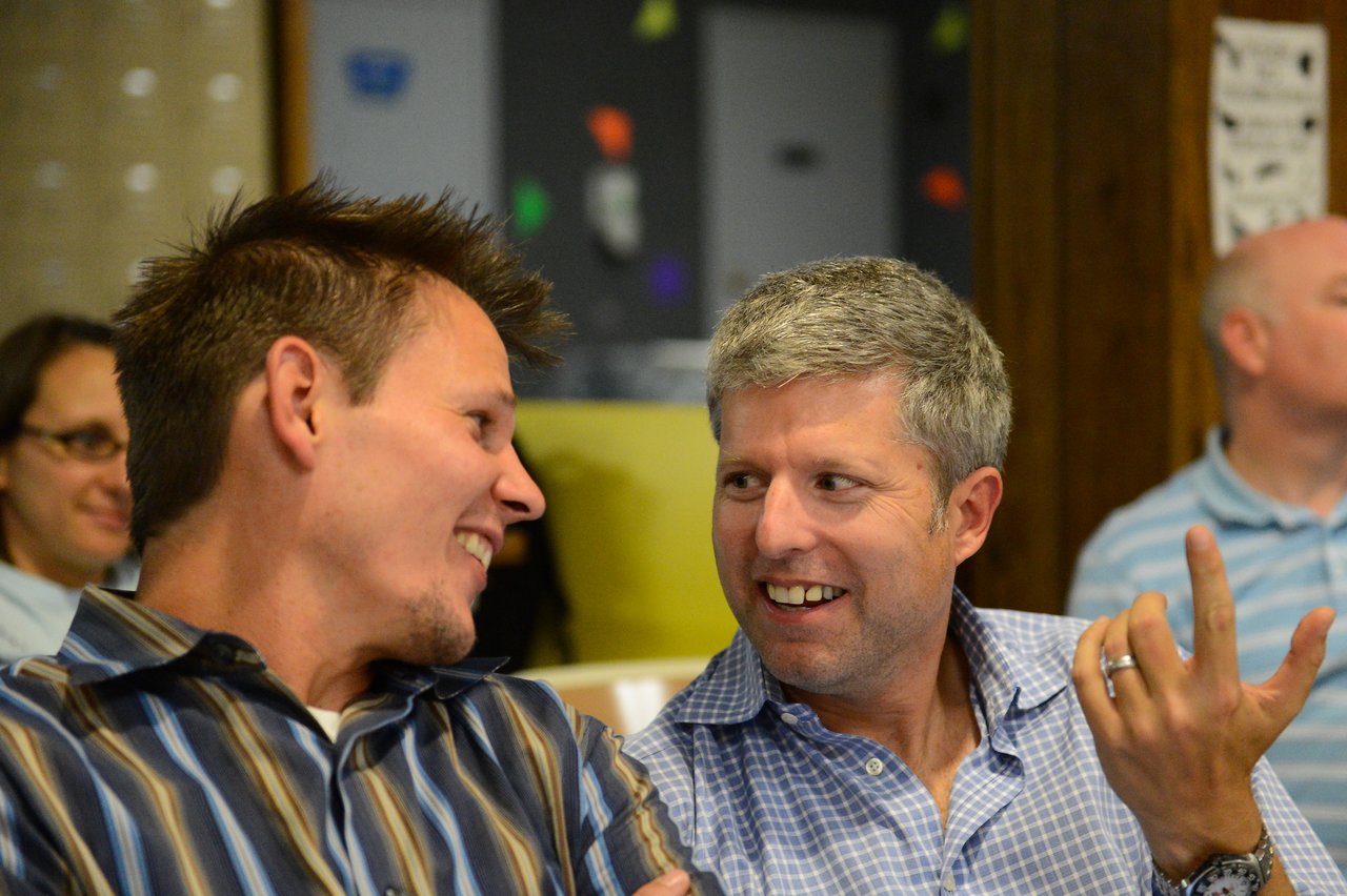 Two men in conversation, smiling and gesturing, while seated in a room with other people in the background.