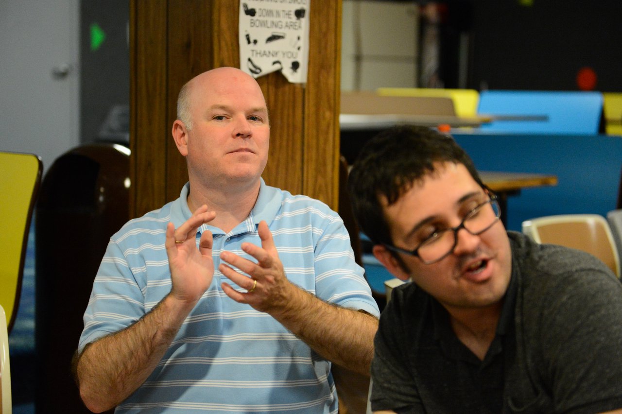 A man in a striped shirt claps while another man in glasses speaks in a casual setting.