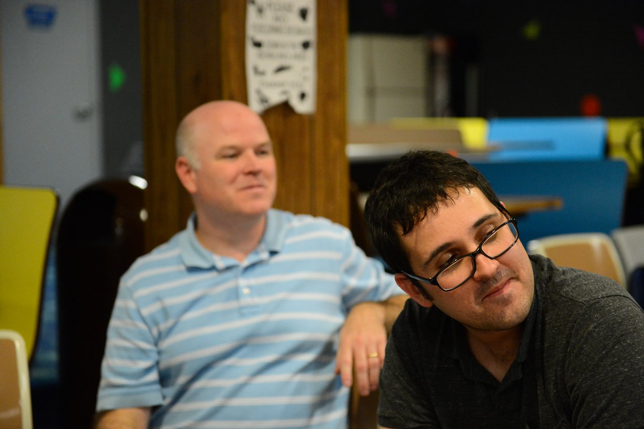 Two men sitting indoors, one in a striped shirt smiling and the other in glasses looking to the side.