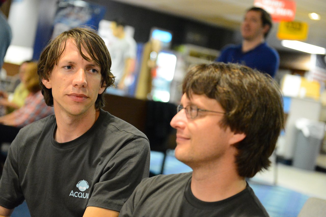 Two people in Acquia shirts sit and talk, while another person stands in the background.