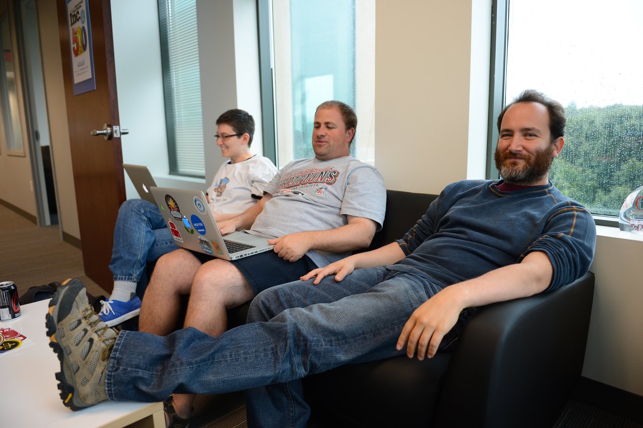 Three people sitting in an office lounge, two using laptops while one relaxes with feet on a table.