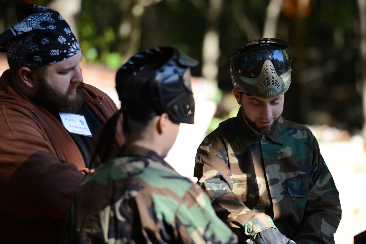 Three people in camouflage and protective gear prepare for a paintball game, with one adjusting equipment.