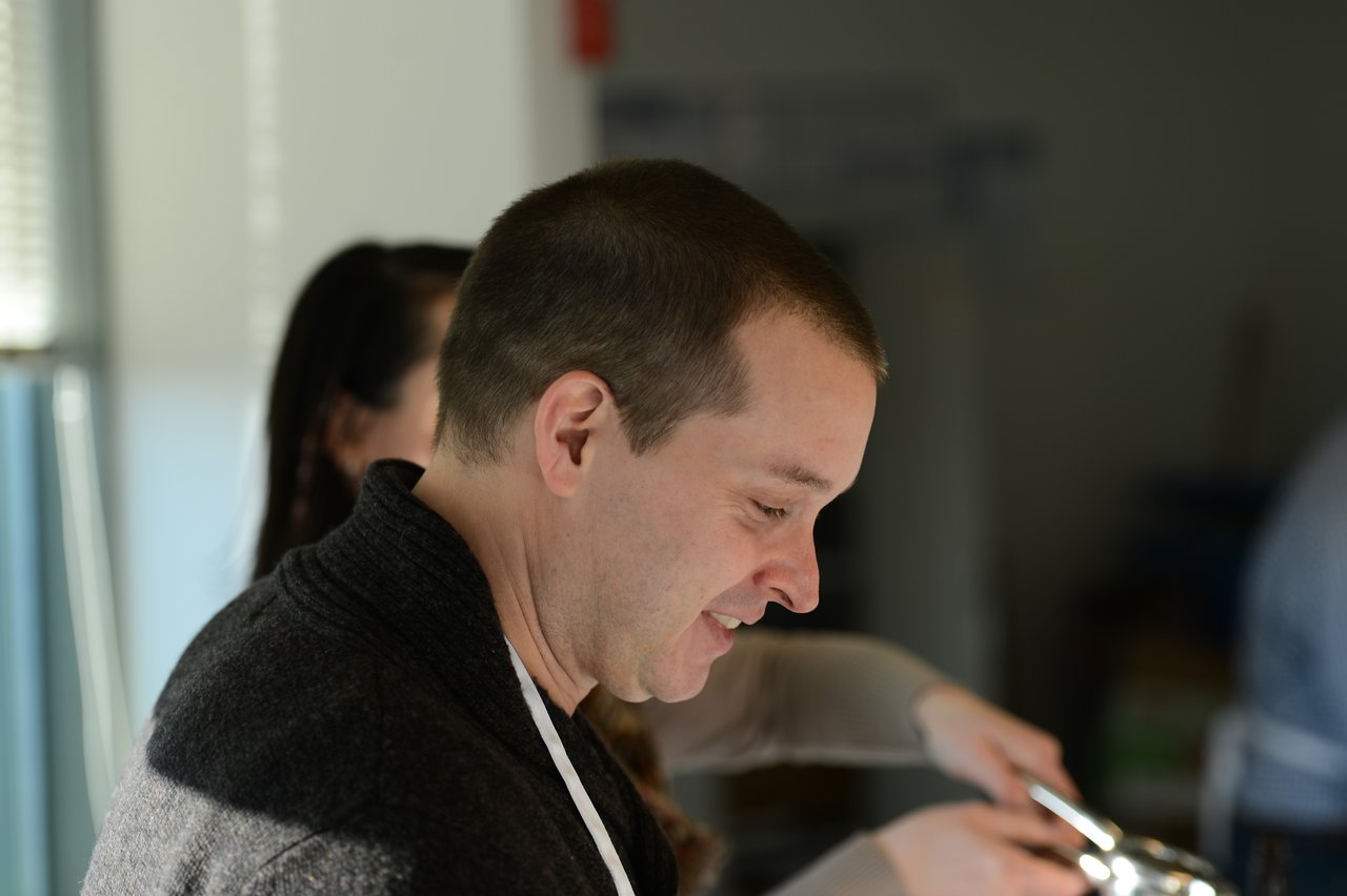 A man in a dark sweater smiles while serving food with tongs at an event.