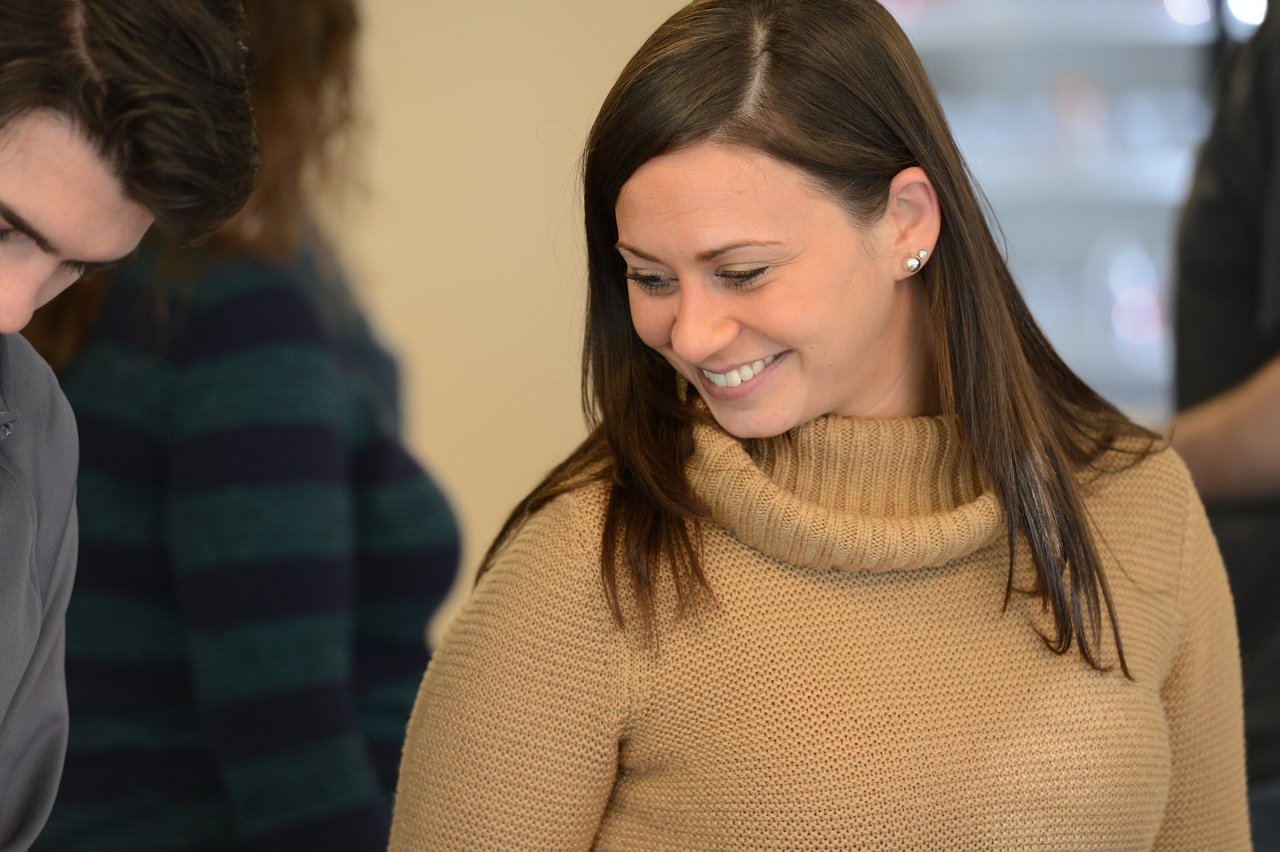 A woman in a beige sweater smiles while looking down, surrounded by other people at an event.