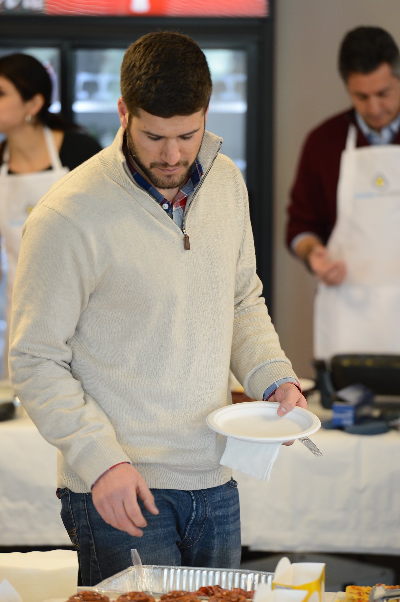 A man in a beige sweater holds a plate while selecting food from a buffet table at an event.