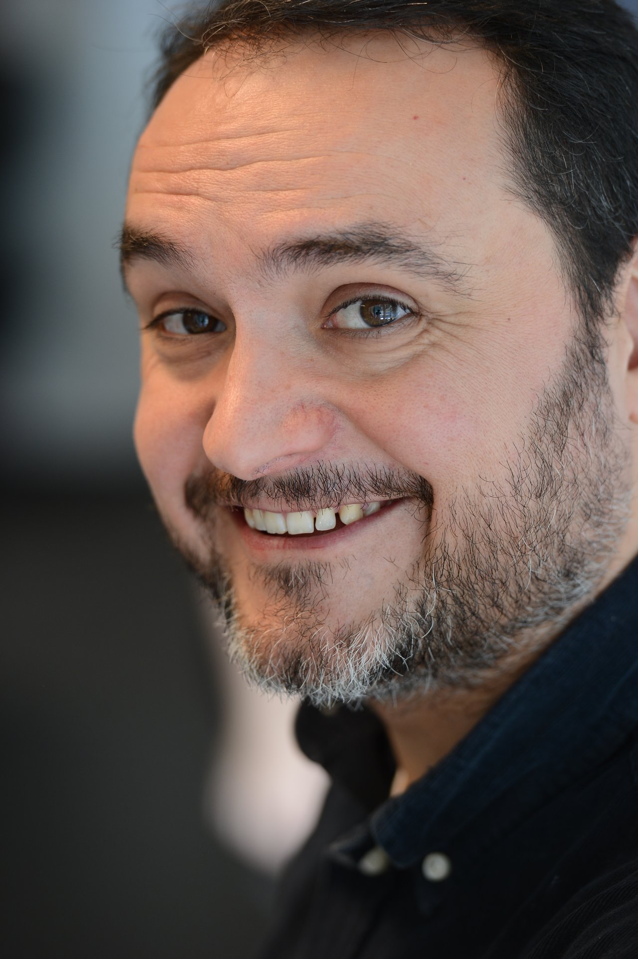 A man with a beard and dark shirt smiles at the camera in a close-up portrait.