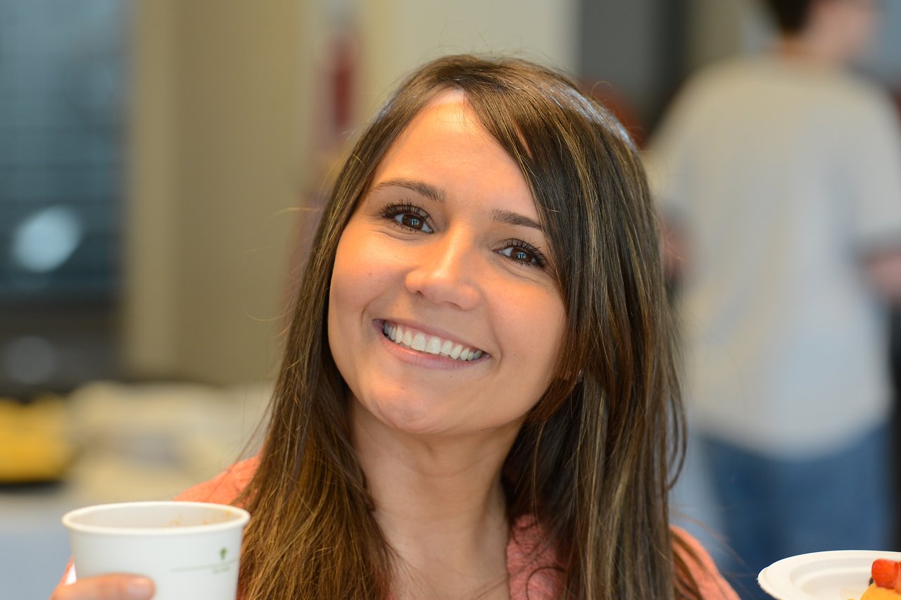 A smiling woman holds a cup and plate at a breakfast event, with other attendees in the background.