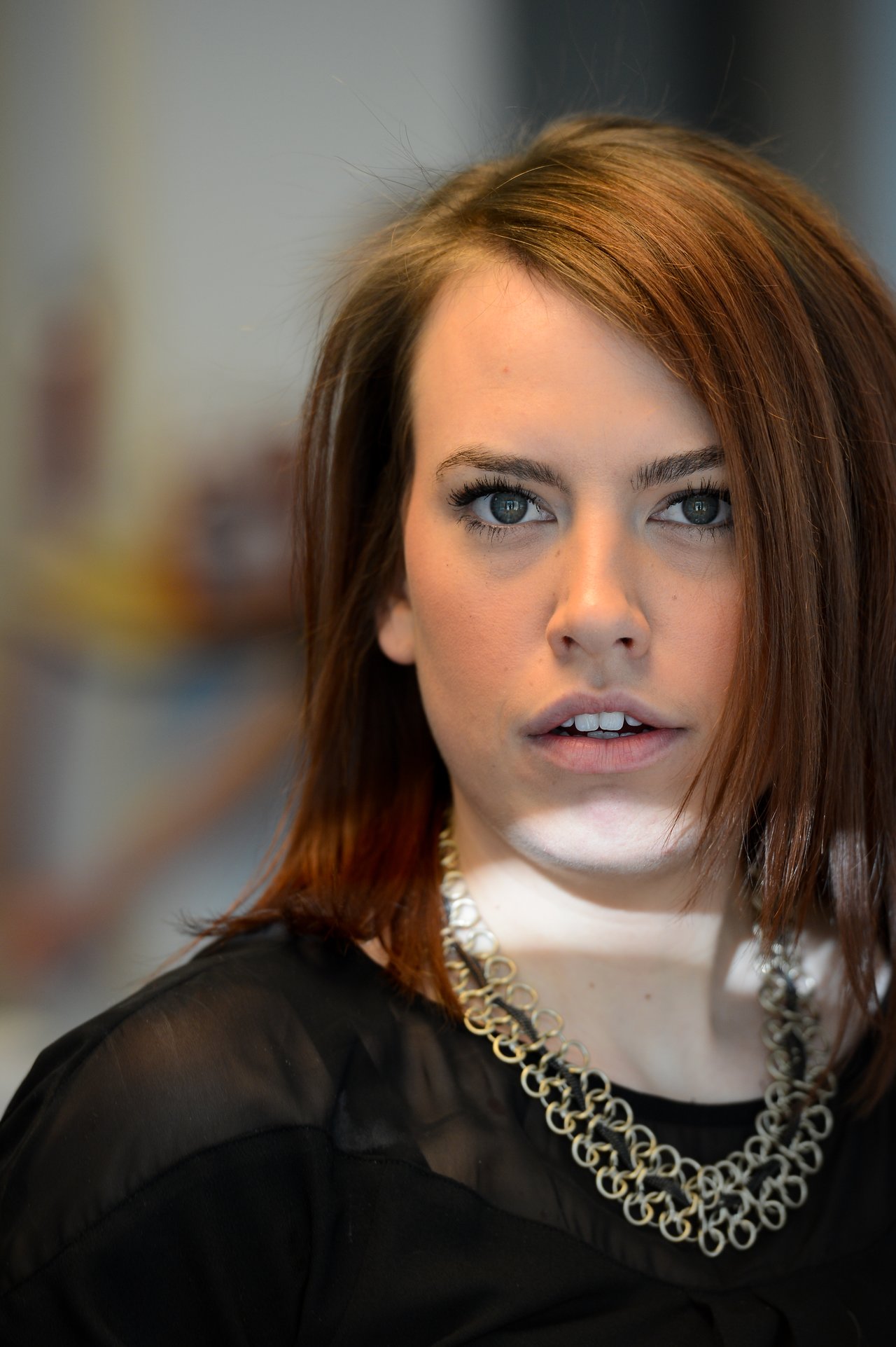 A woman with shoulder-length brown hair and a black outfit looks directly at the camera with a neutral expression.