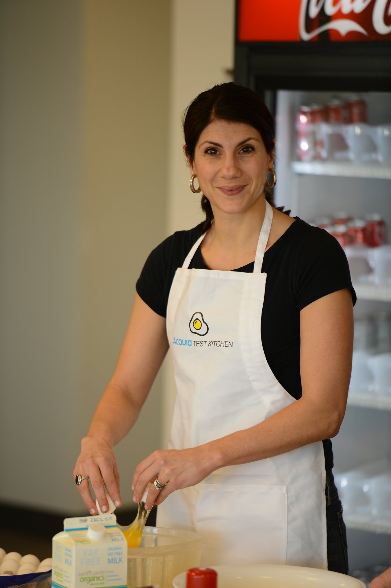 A woman wearing an "Acquia Test Kitchen" apron cracks an egg into a bowl while preparing food.