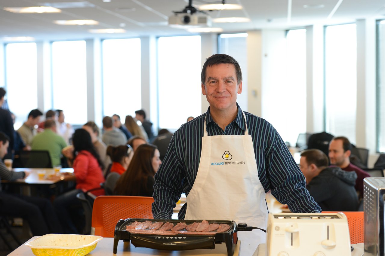 A man wearing an "Acquia Test Kitchen" apron cooks bacon on a griddle at a breakfast event.