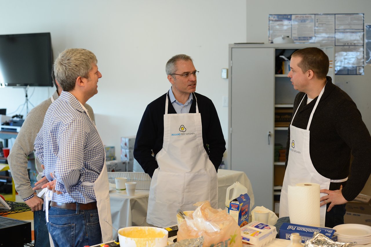 A group of people wearing aprons stand around a table, engaged in conversation at a cooking event.