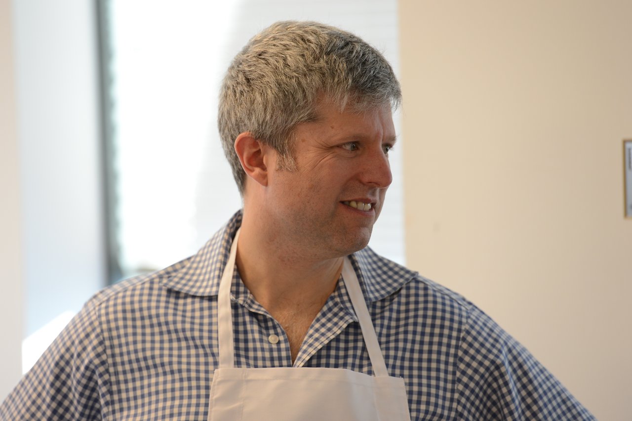 A man in a checkered shirt and apron smiles while looking to the side at an indoor event.