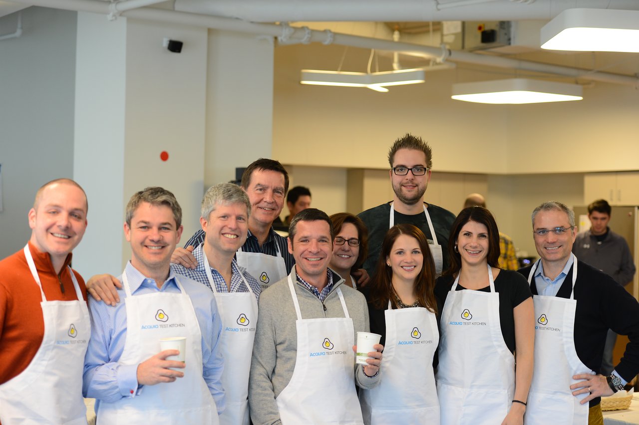 A group of people wearing Acquia-branded aprons pose together in a kitchen, smiling and holding cups.