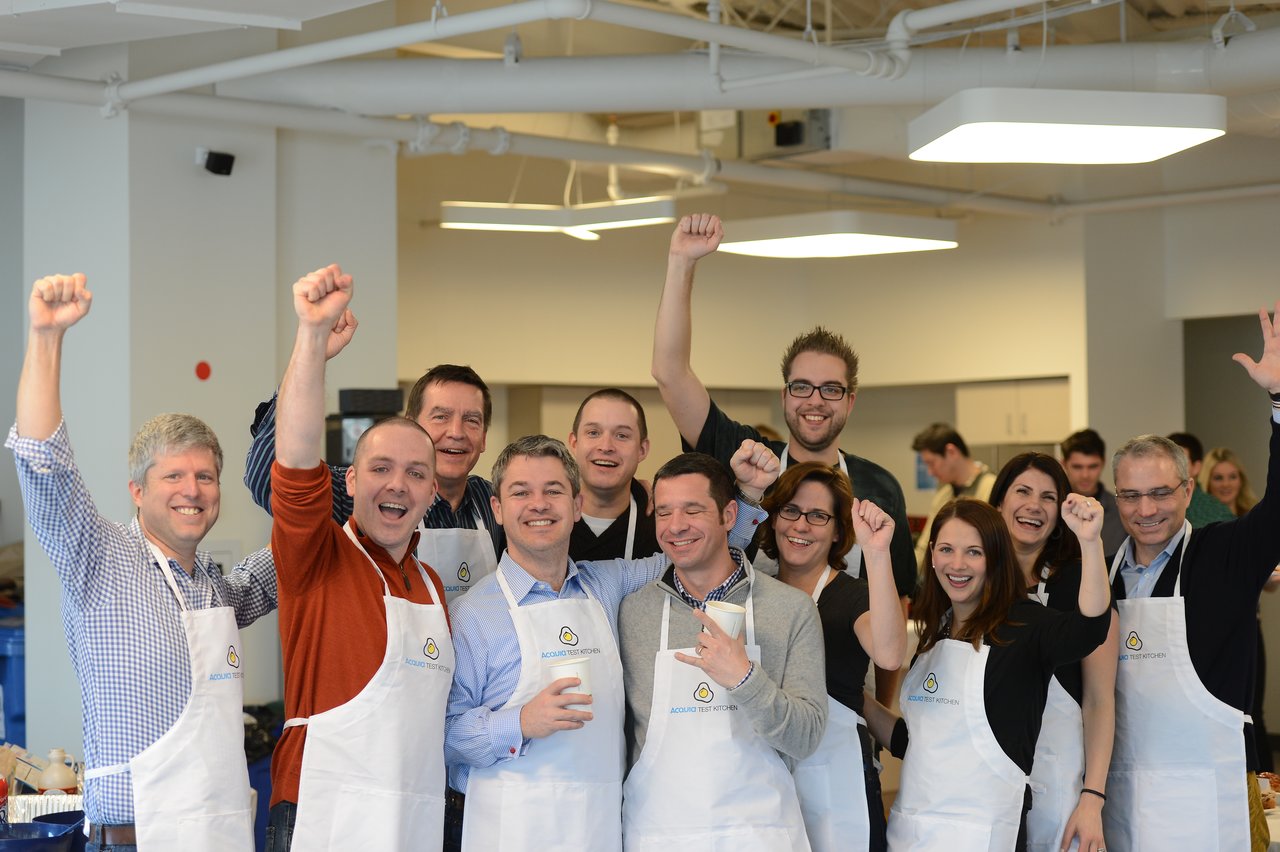 A group of people wearing aprons cheer and raise their fists at an Acquia executive breakfast event.