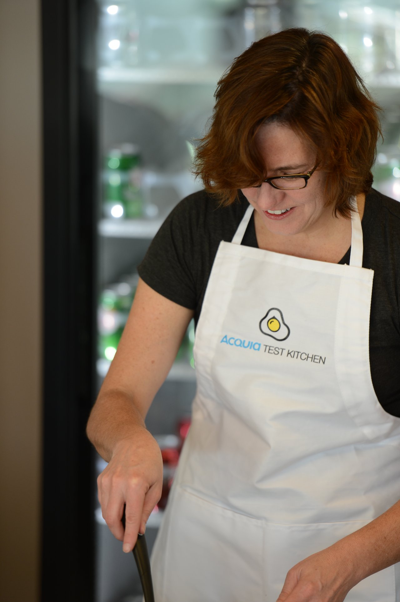 A person wearing an "Acquia Test Kitchen" apron is cooking and smiling while looking down at their task.