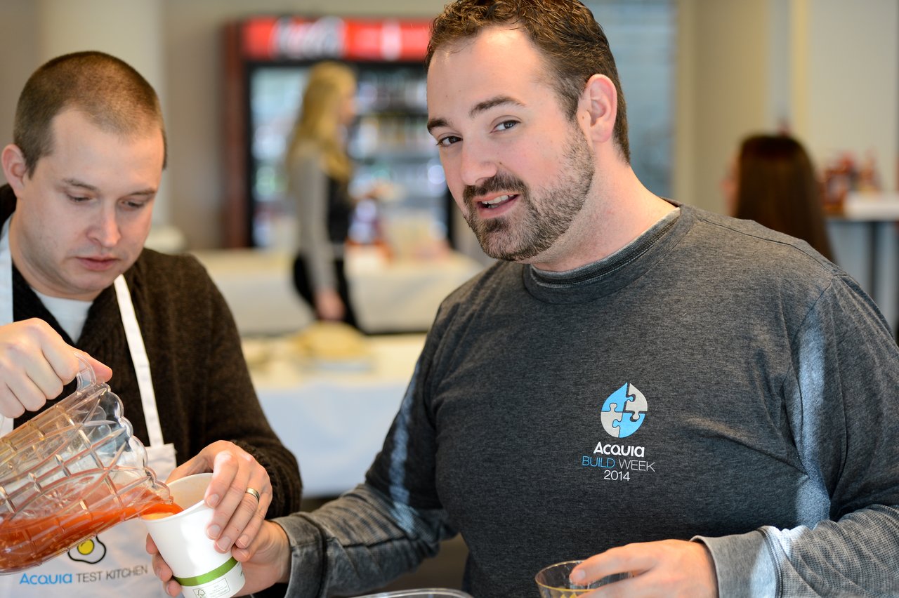 A person pours a red beverage into a cup while another person in an Acquia shirt looks toward the camera.