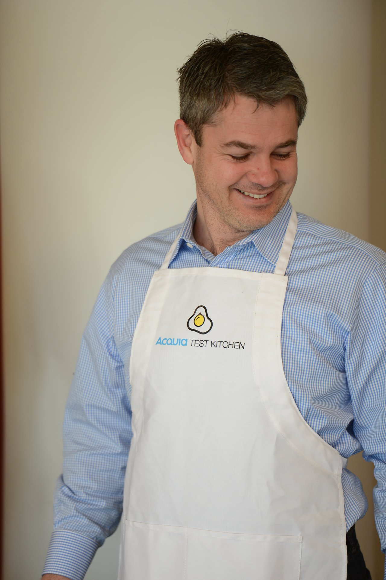 A man wearing a white "Acquia Test Kitchen" apron smiles while looking down.