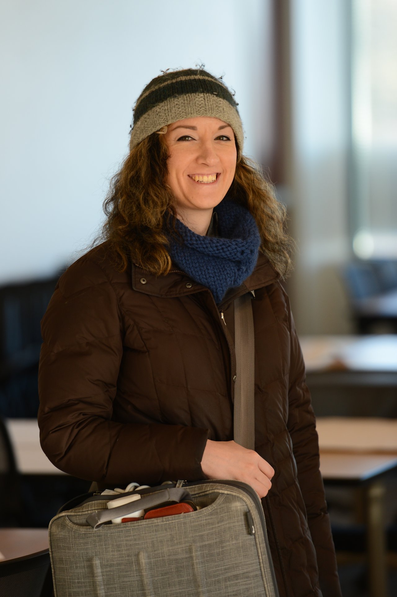 A person wearing a winter coat and hat smiles while carrying a shoulder bag in an indoor setting.