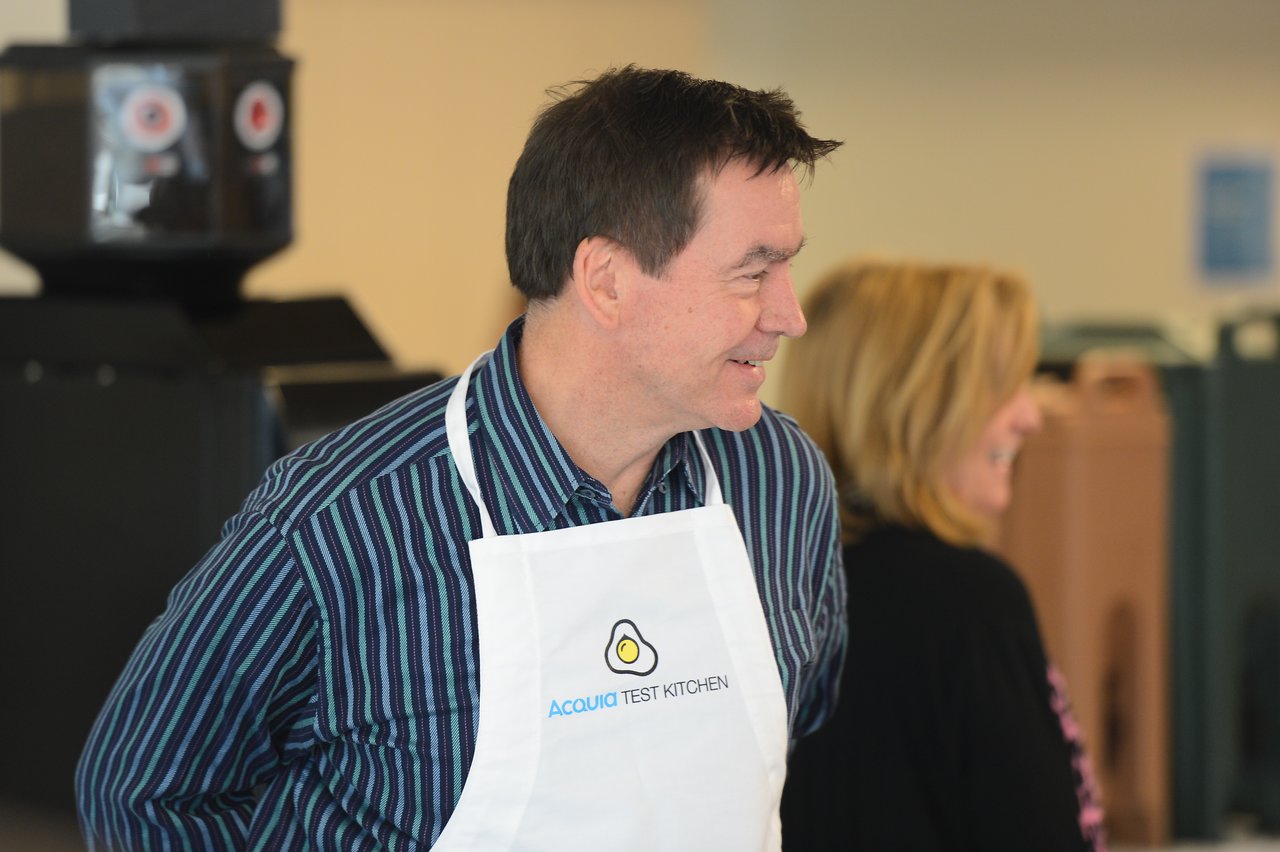 A man wearing an "Acquia Test Kitchen" apron smiles while engaging in conversation at an event.
