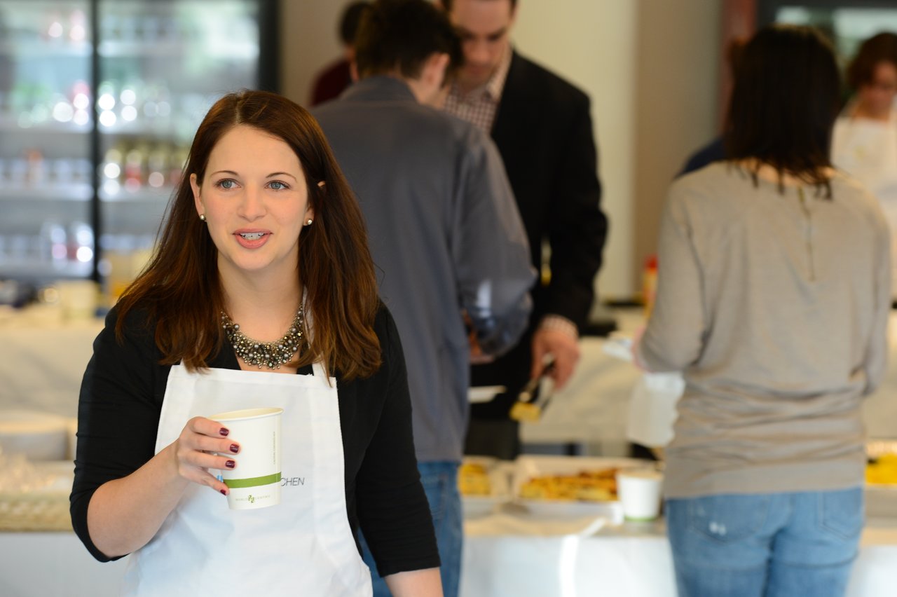 A woman in a white apron holds a cup and speaks, while others serve themselves food in the background.