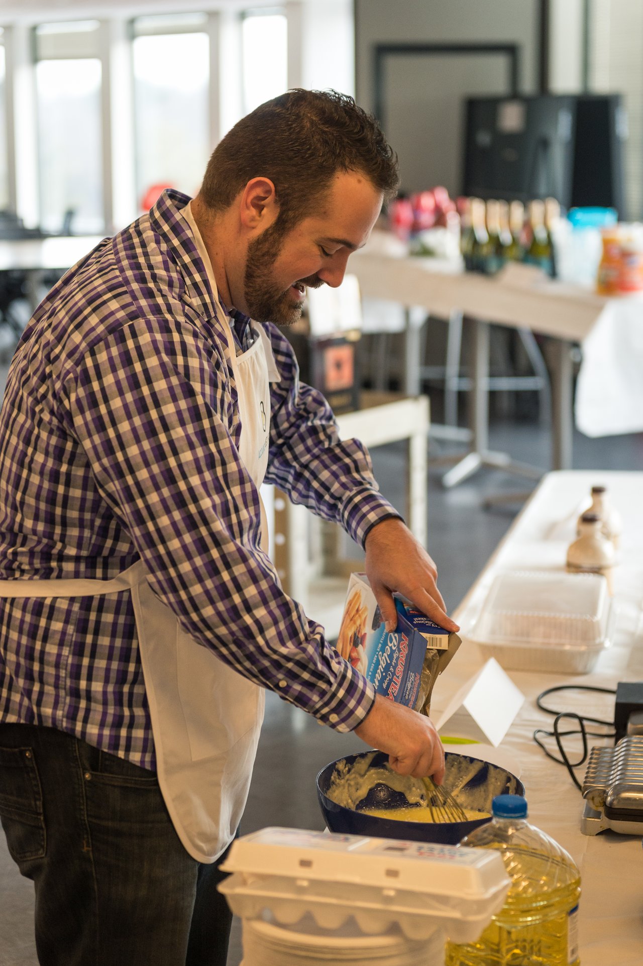 A man wearing an apron mixes batter in a bowl while pouring ingredients from a box at a breakfast event.