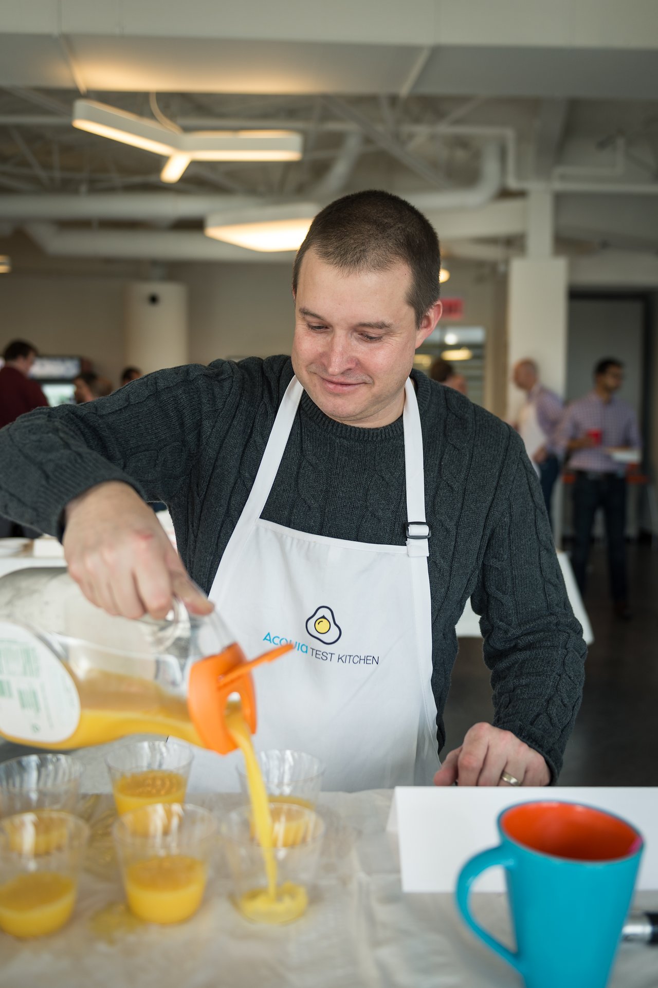 A person wearing an "Acquia Test Kitchen" apron pours orange juice into plastic cups at a breakfast event.
