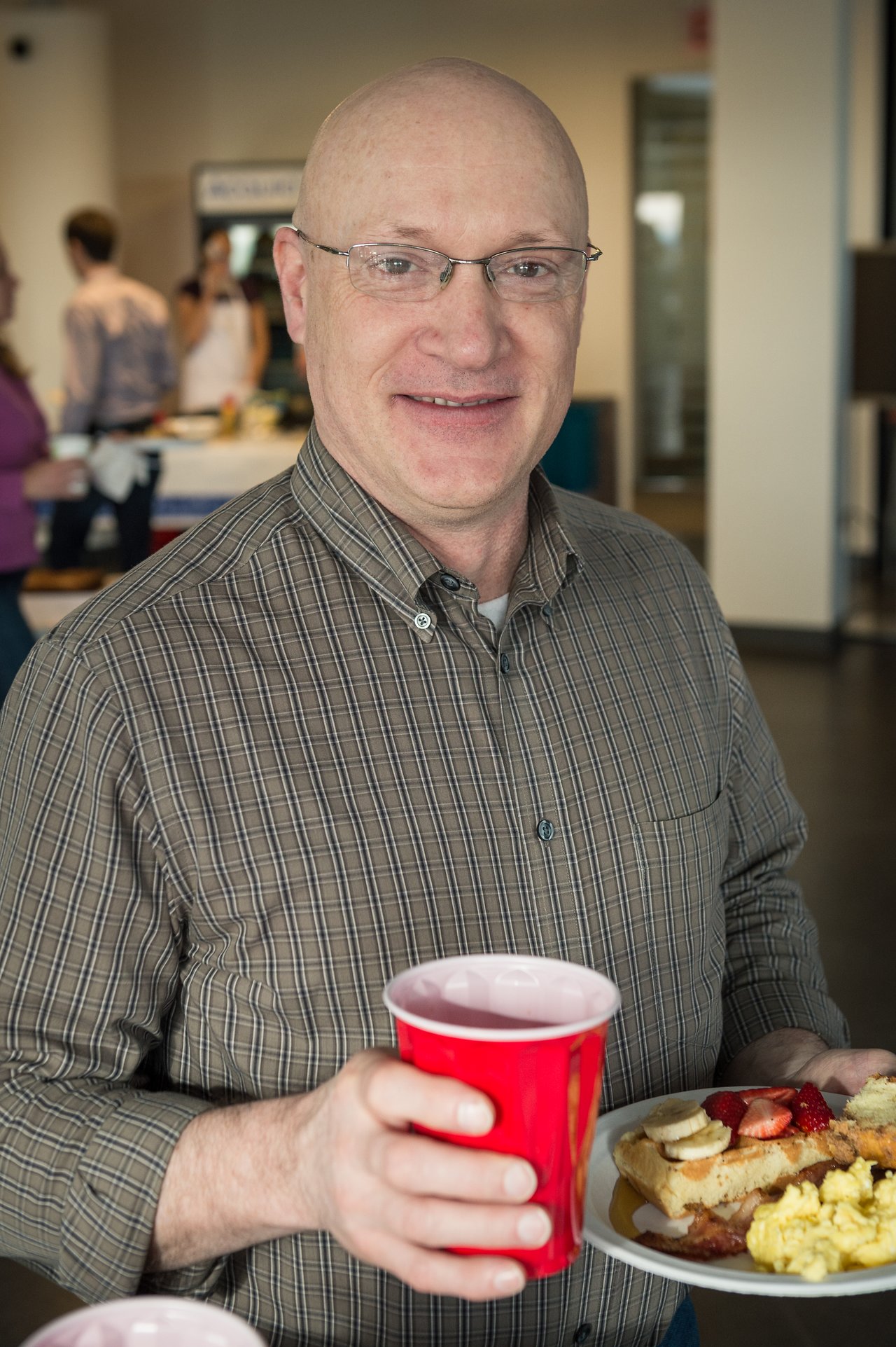 A man in a plaid shirt holds a red cup and a plate of breakfast food, smiling at the camera.