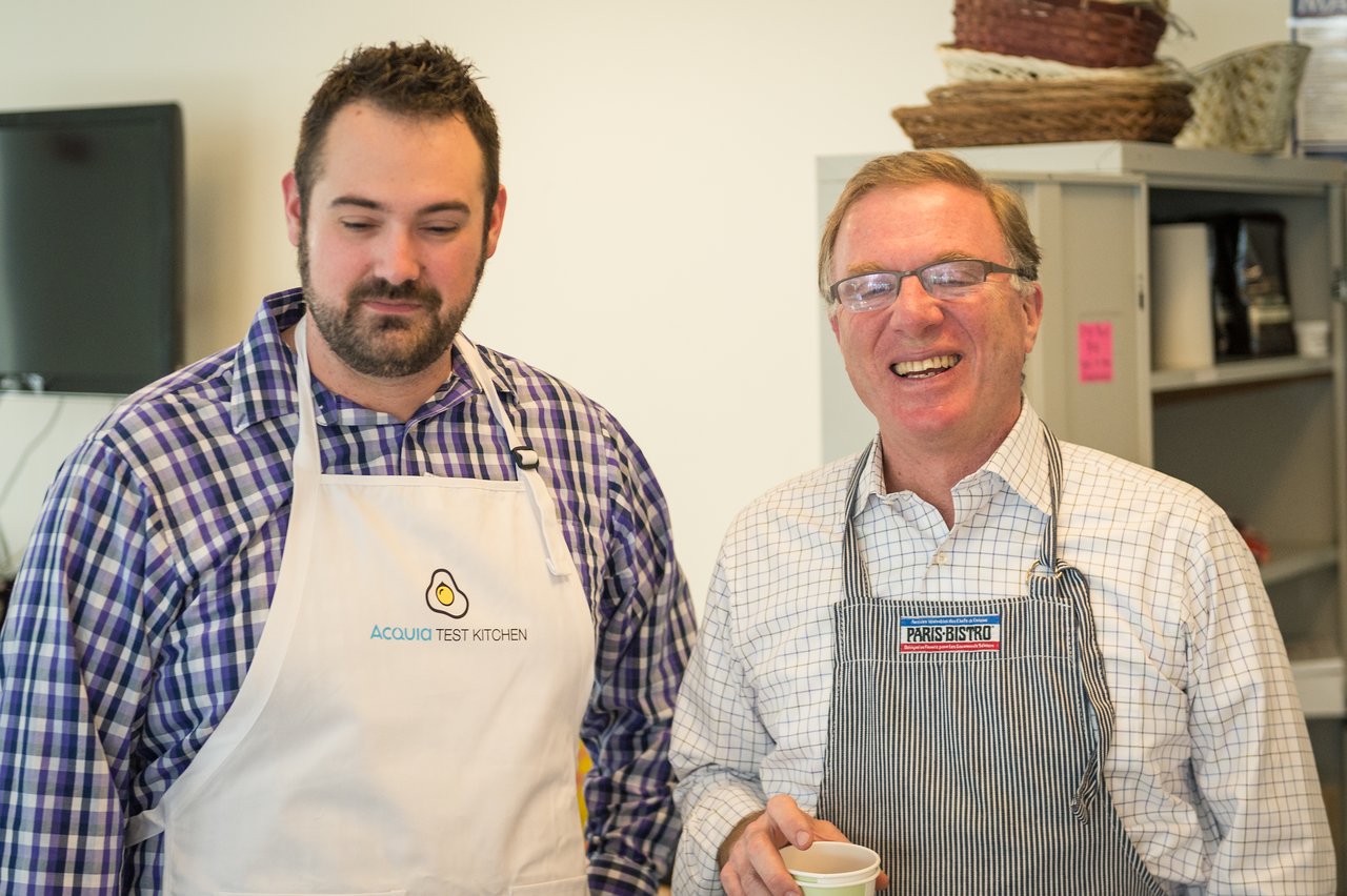 Two men wearing aprons stand together, one smiling and holding a cup, at an Acquia event.