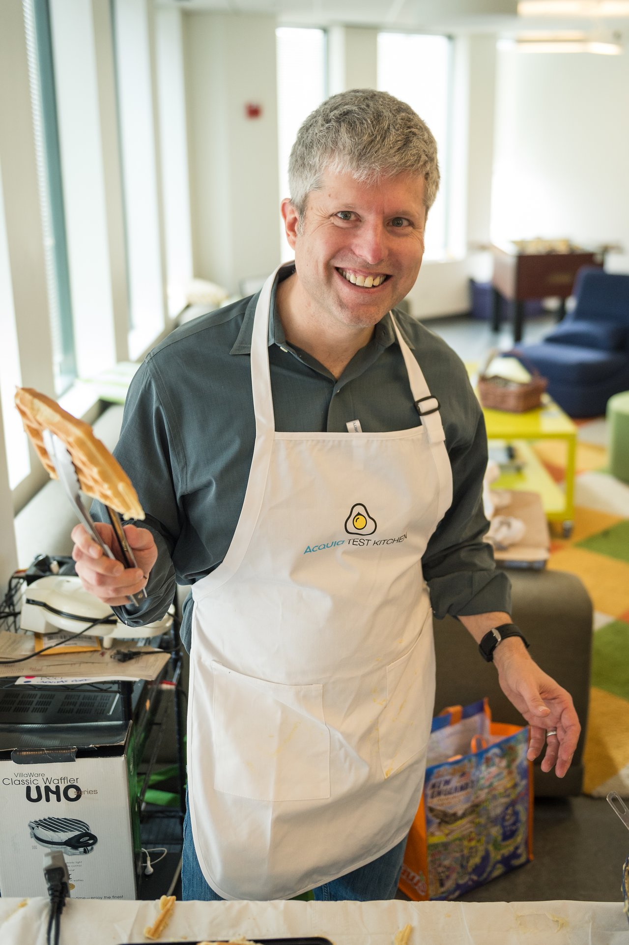 A smiling person wearing an apron holds tongs with a waffle, standing behind a waffle-making station.
