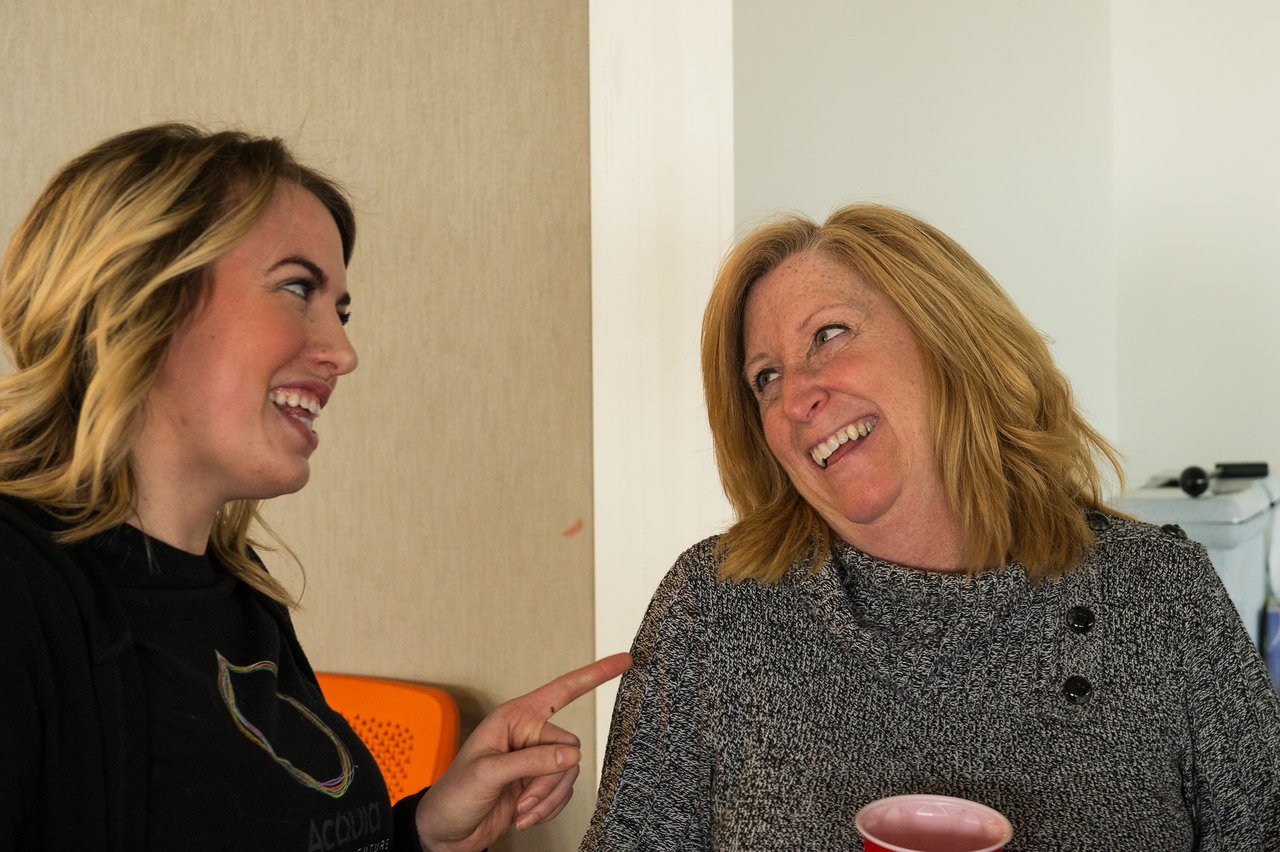 Two women smiling and talking, one pointing while the other holds a cup, at an Acquia event.