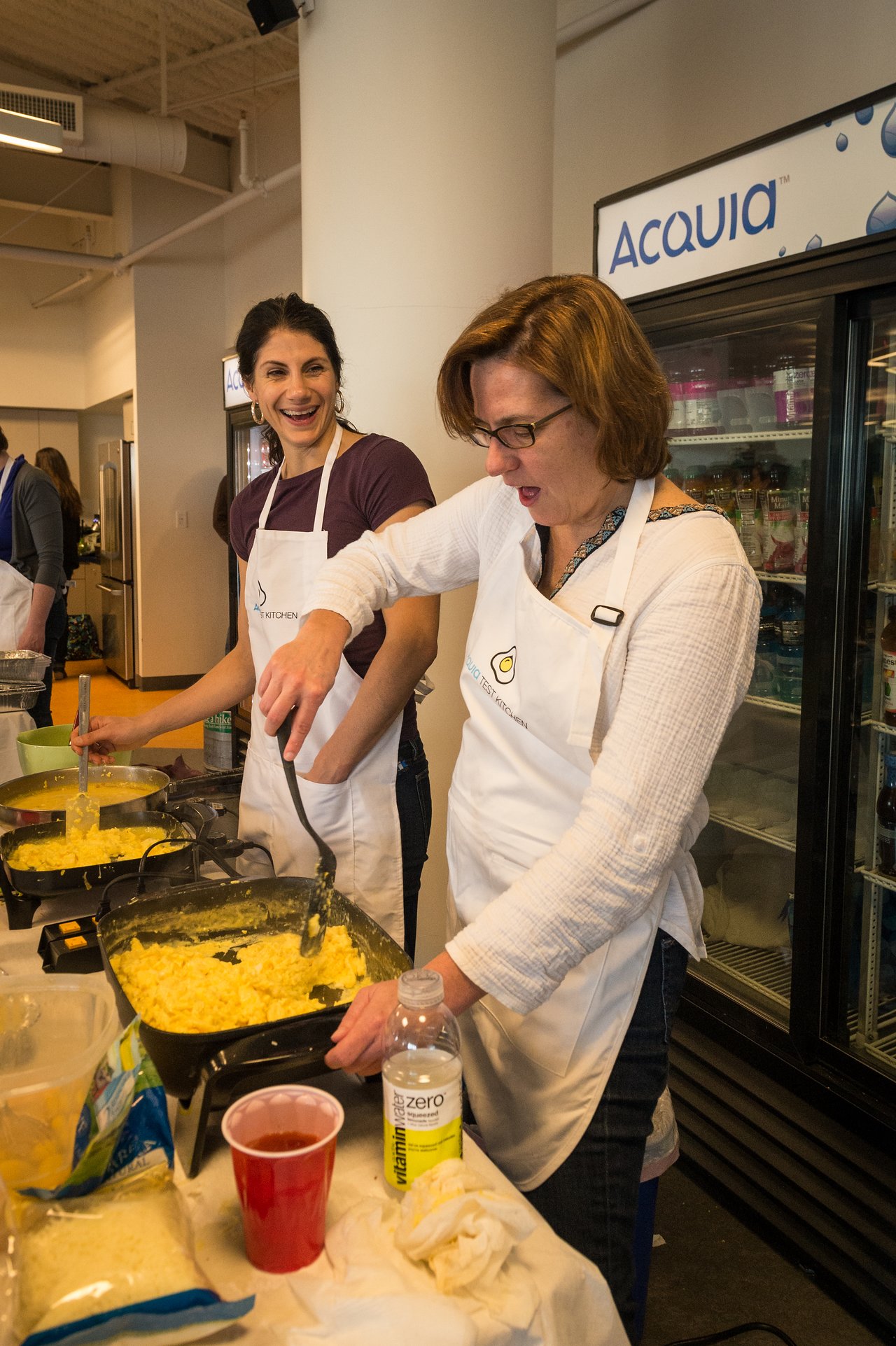 Two people wearing aprons cook scrambled eggs on a griddle, smiling and engaged in conversation at an Acquia event.