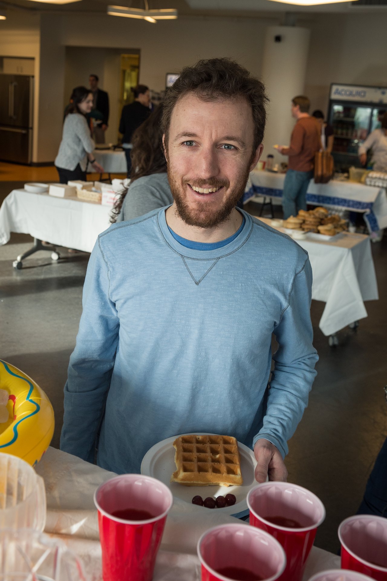 A man in a blue shirt smiles while holding a plate with a waffle and grapes at a breakfast event.