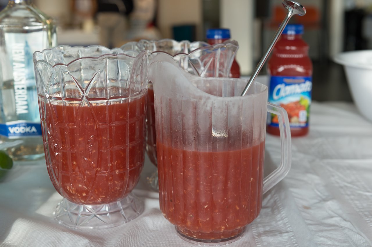 Two pitchers filled with red tomato-based drink on a table, with a stirring spoon and bottles in the background.